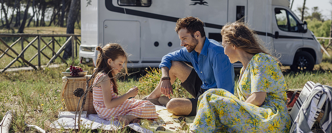 Eltern mit Kind sitzen auf Picknickdecke, parkendes Wohnmobil im Hintergrund